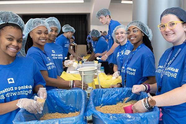 A diverse group of students smiles at the camera while packing food at a volunteer event. They are all wearing hairnets and shirts that say “Making a Difference Begins With Blue.” Other students are visible working in the background.