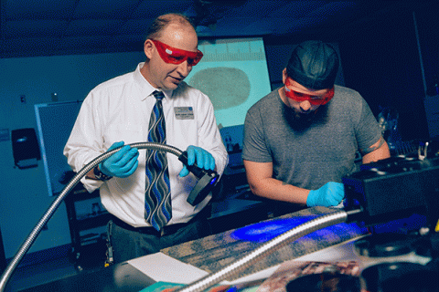 A male criminology professor with a white shirt and tie and red safety glasses shines a blue light on a sample as a student looks on with a projection of a fingerprint in the background.